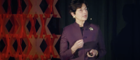 Woman in purple jacket gestures while speaking in front of a dark patterned backdrop